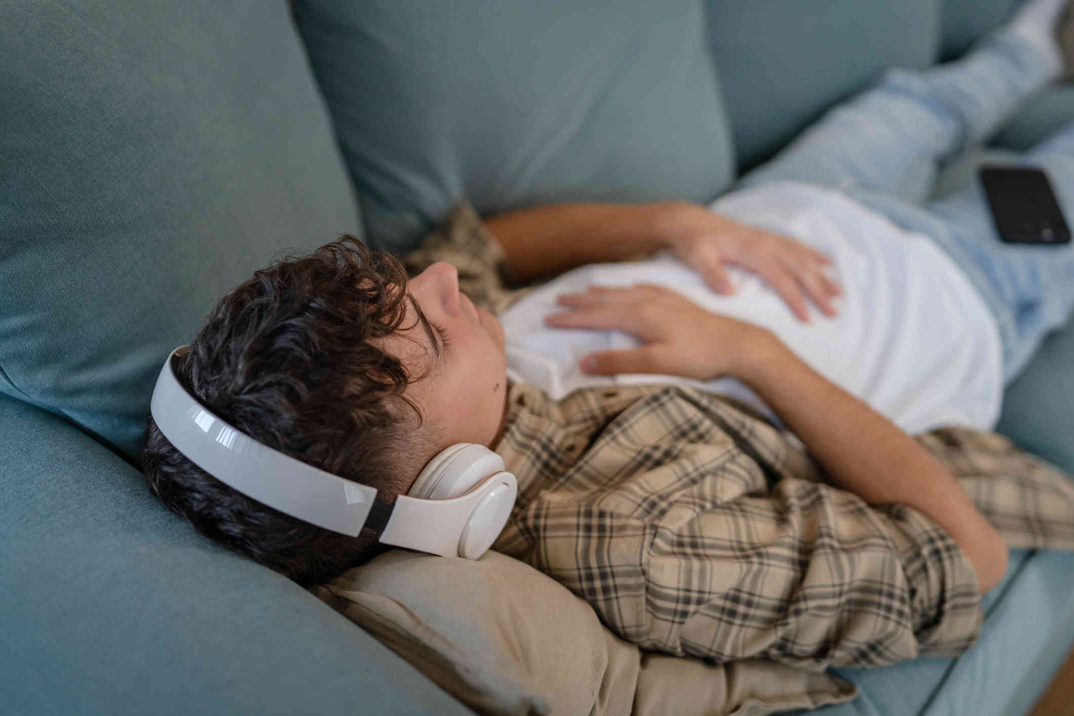 A young man in a plaid flannel lays on the couch with headphones on and eyes closed while hands resting on chest